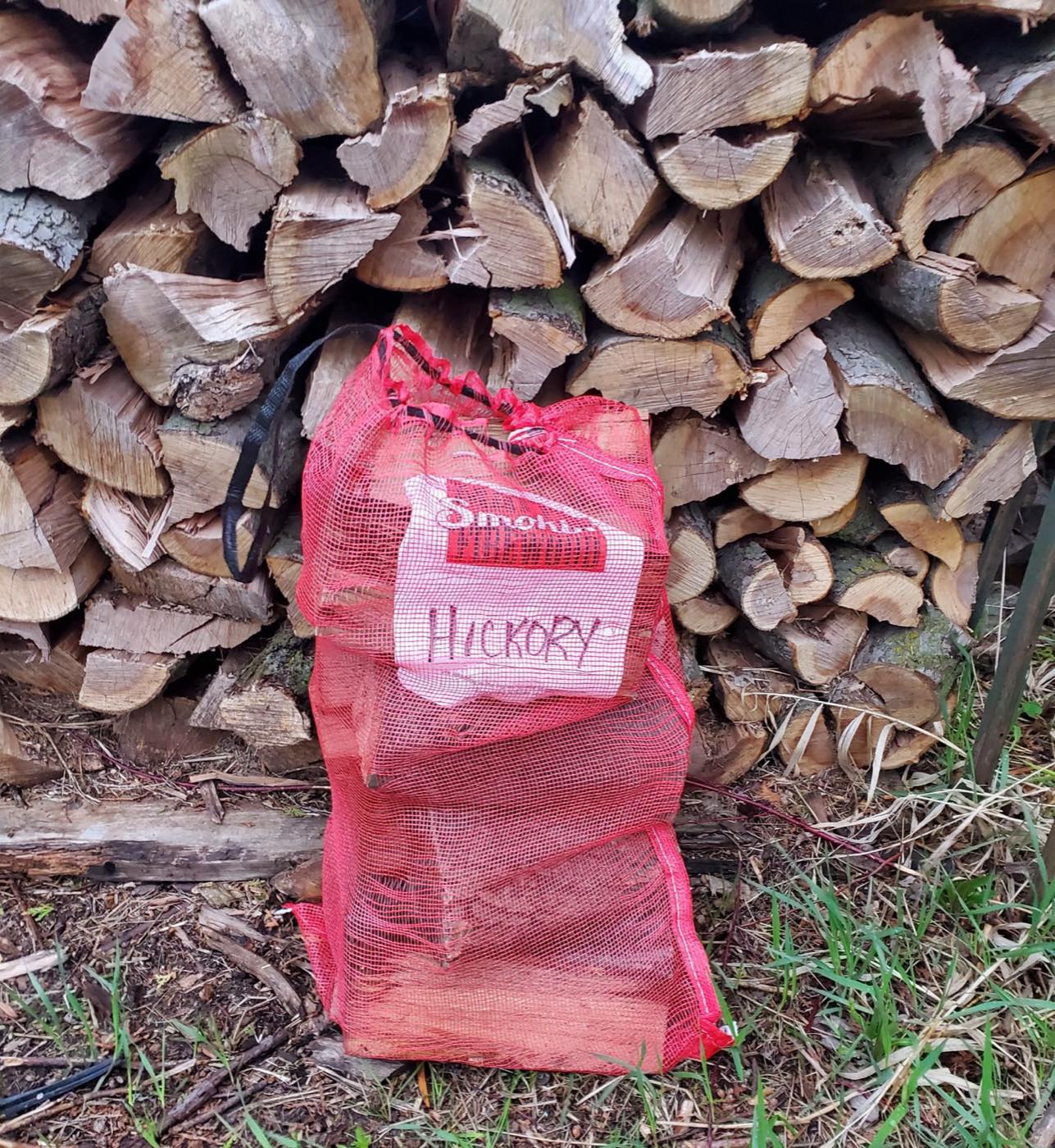Red mesh bag of hardwood firewood standing in front of a stacked woodpile