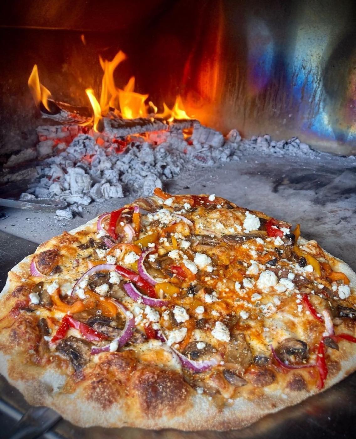 Wood-fired pizza cooking beside a live flame in a stone pizza oven